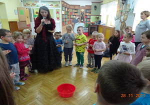 Dzieci stoją wokół nauczycielki przebranej za Cygankę. Na środku stoi czerwona miska z wodą.